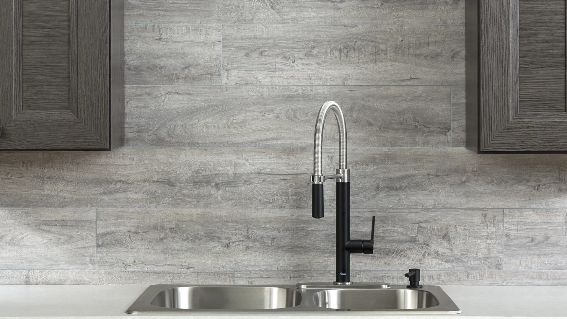 Modern kitchen sink with dual stainless steel basins, black and chrome faucet, gray wood cabinets, and light gray backsplash.
