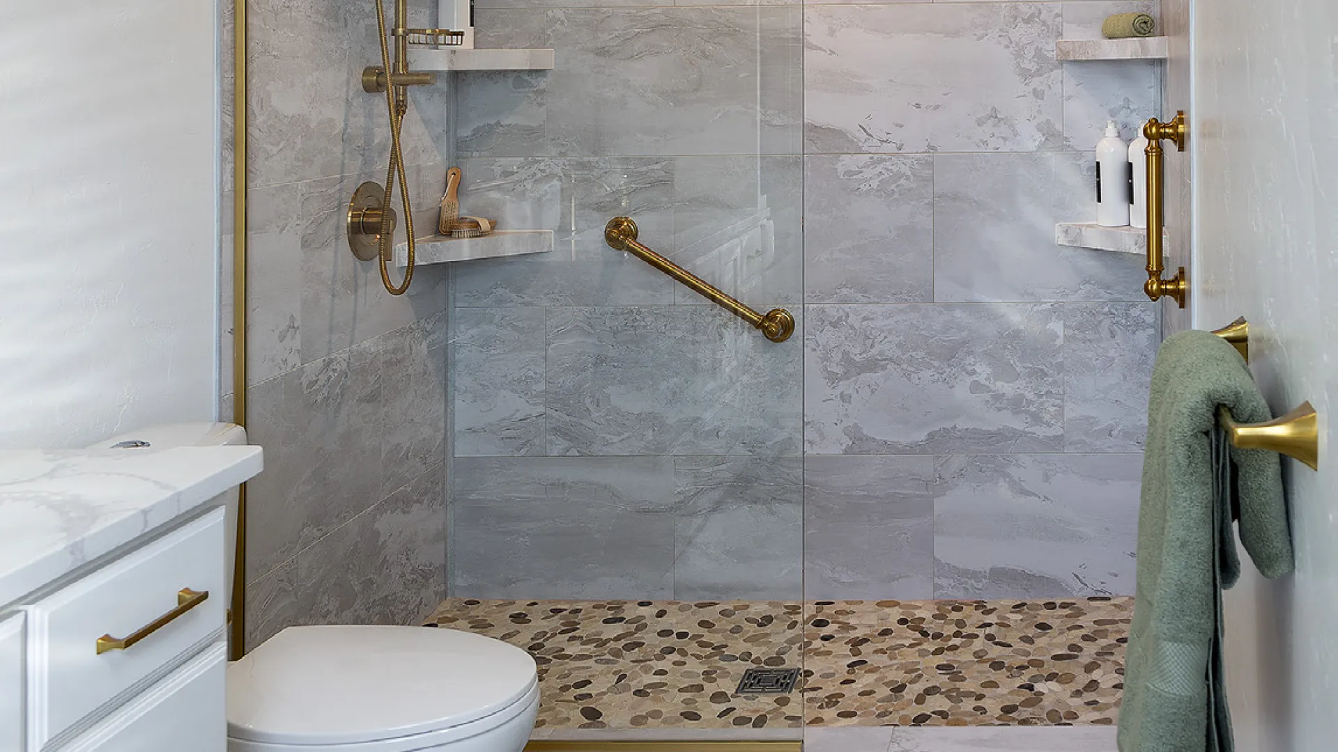 Modern bathroom with stone tile shower, brass fixtures, white toilet, gray floor, and green towel on brass hook.