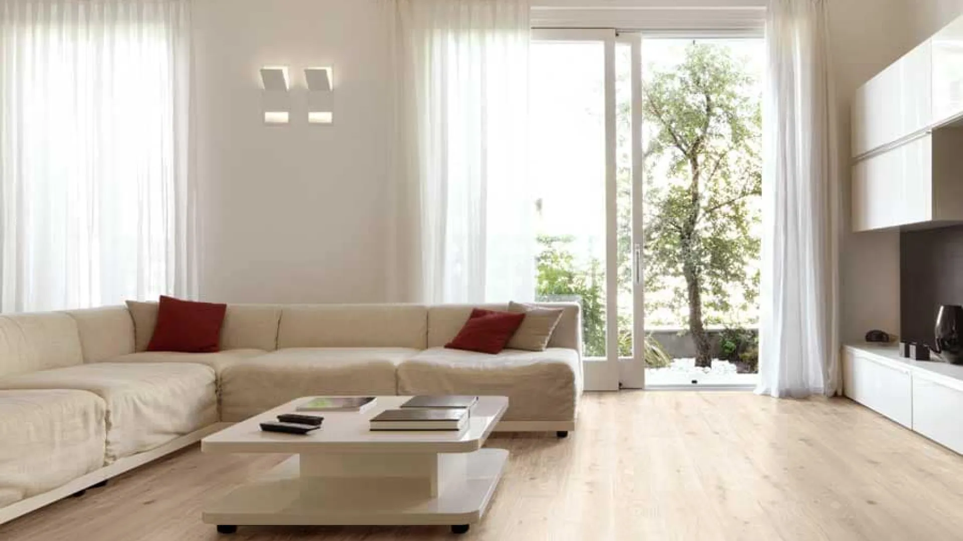 Bright modern living room with large beige sectional sofa, white coffee table, light wood flooring, and large windows.