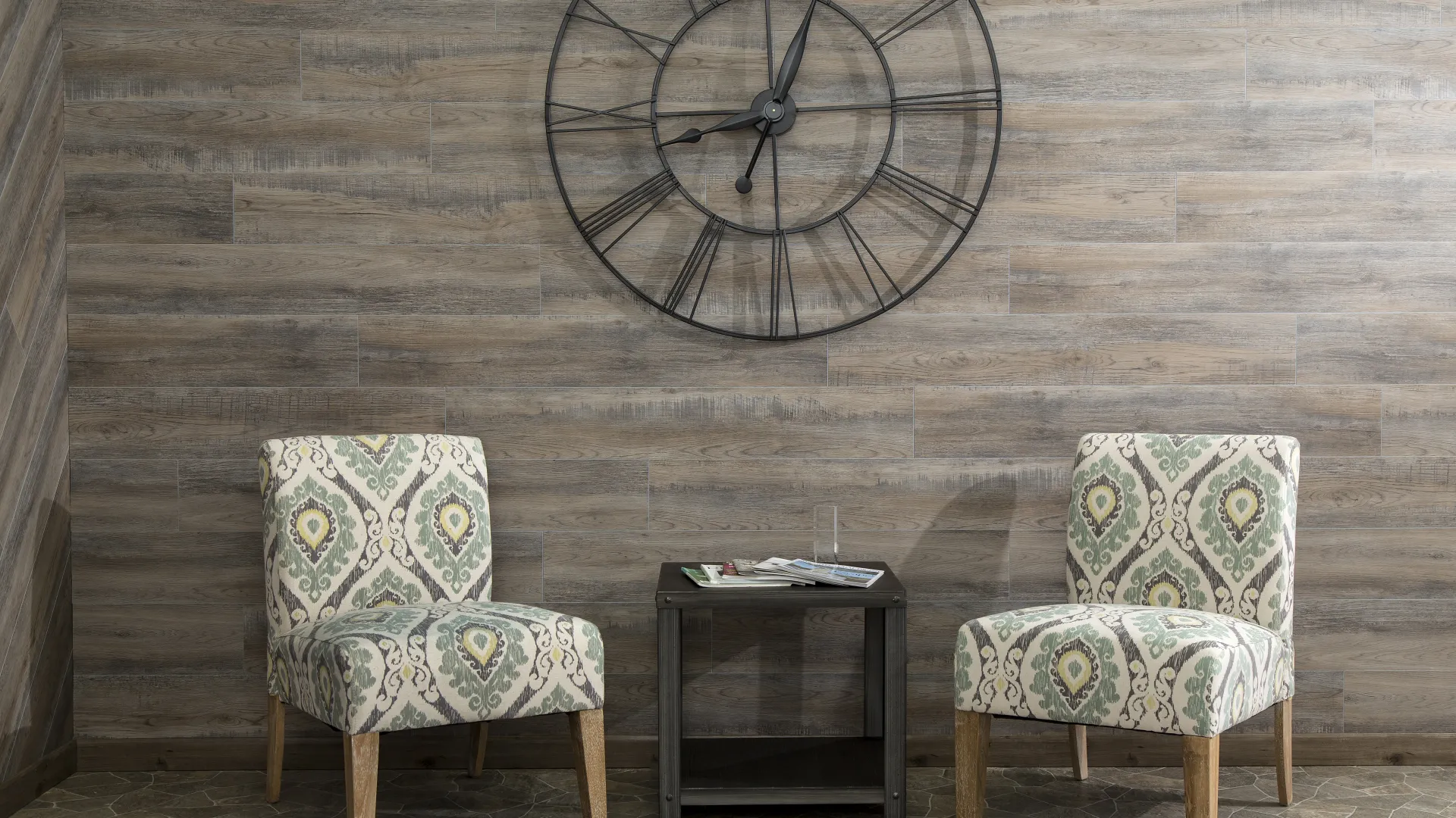 Two patterned armchairs with wooden legs and a small black table below a large black metal wall clock on a wood-paneled wall.