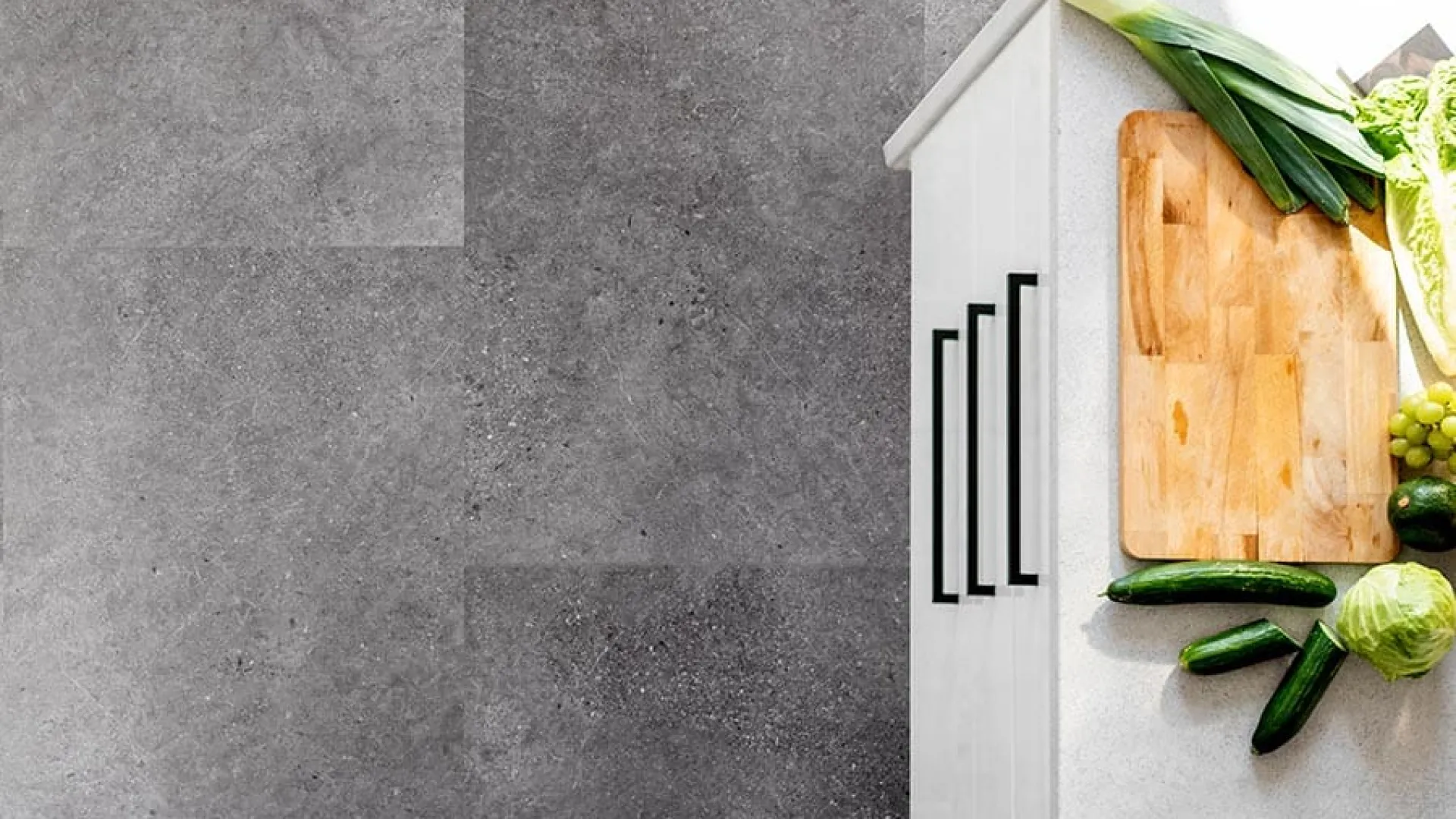 Top view of modern kitchen countertop with vegetables, cutting board, knife, and white cabinets with black handles.