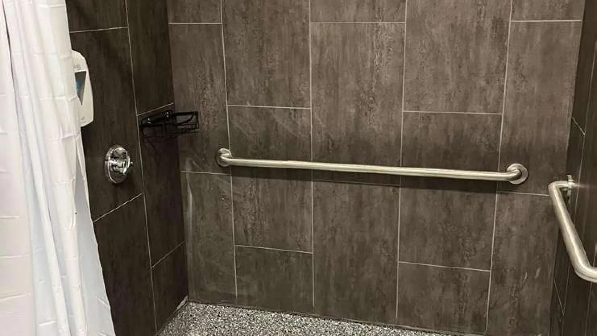 Accessible walk-in shower with gray tiled walls, grab bars, white shower curtain, and textured floor.