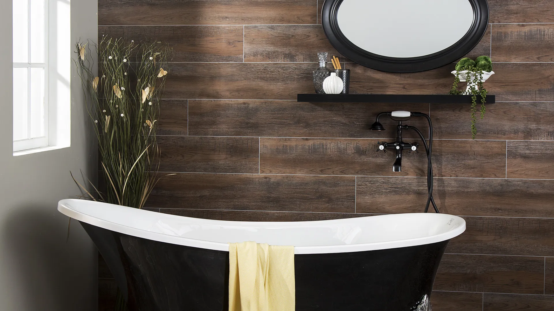 Black clawfoot bathtub with yellow towel, wooden wall tiles, oval mirror, and decorative plants in a bright bathroom.