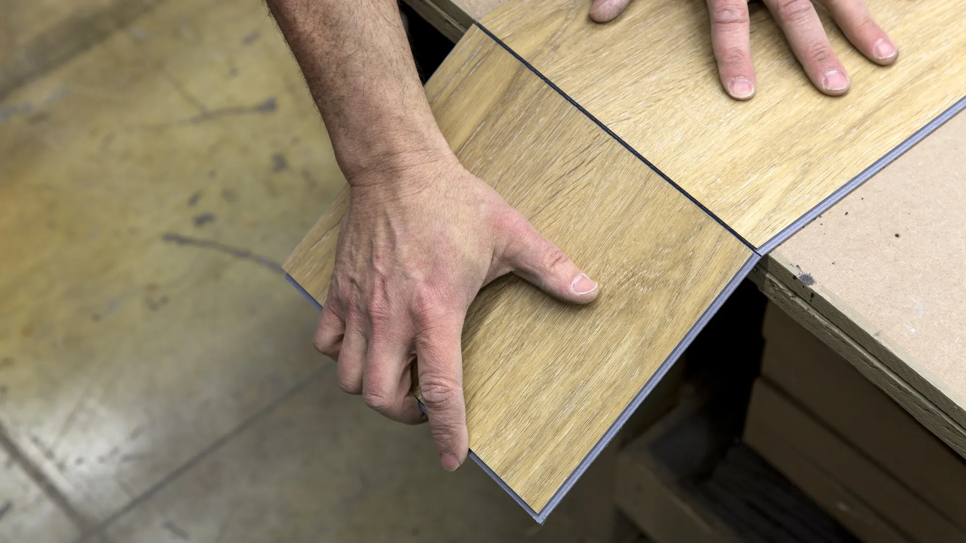 Hands fitting two wood-look laminate flooring planks together on a workshop table.