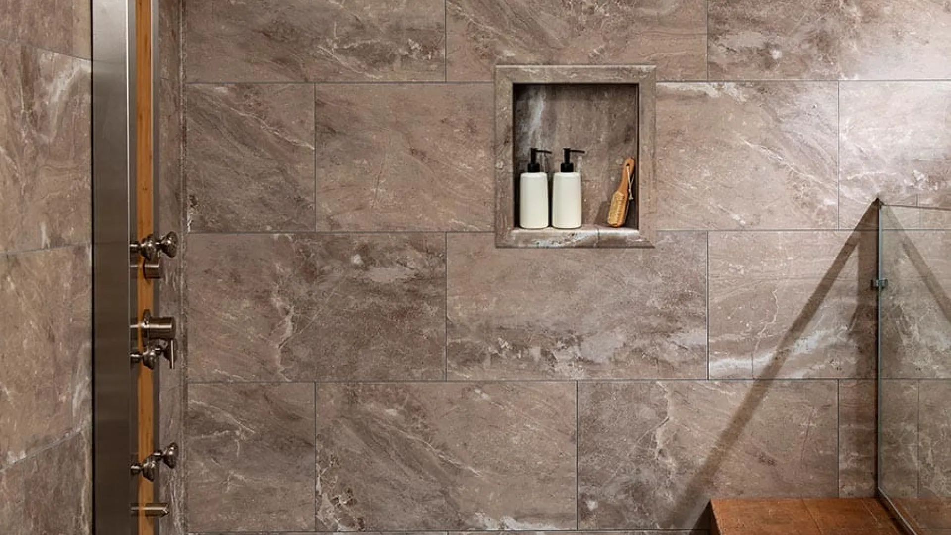 Modern shower area with brown marble tiles, wall-mounted shower panel, built-in shelf, and wooden bench with towels.