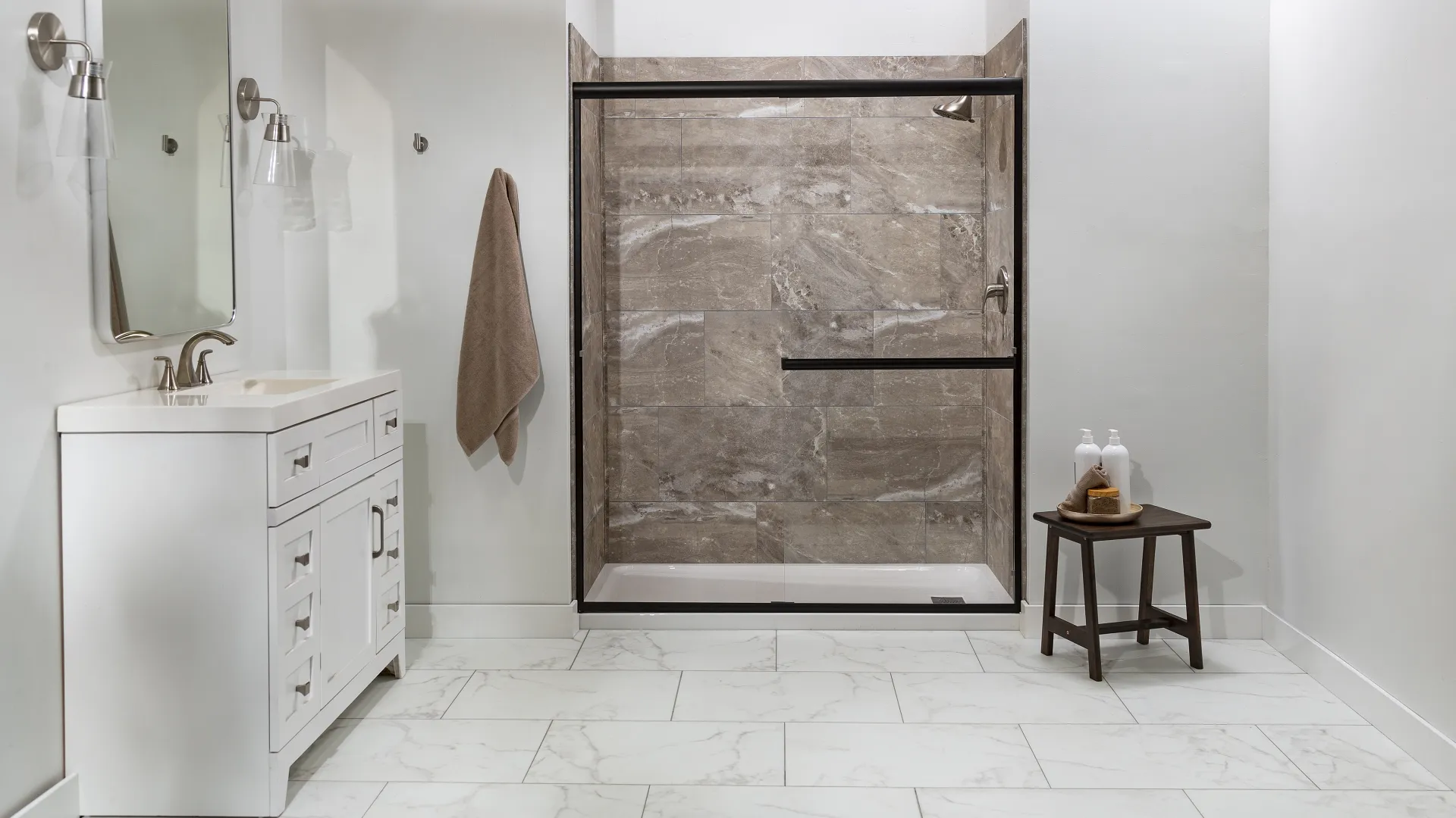 Modern bathroom with white vanity, marble floor, glass shower enclosure, towel, and small wooden stool with toiletries