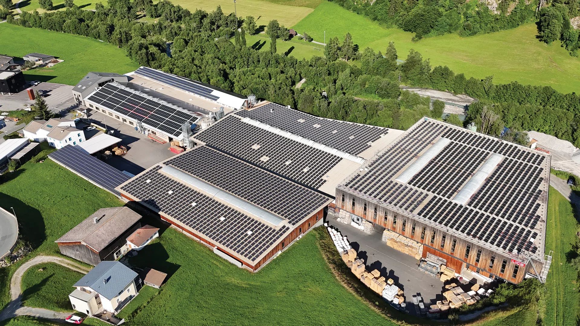Aerial view of industrial buildings with extensive solar panel arrays on rooftops in a green rural landscape.