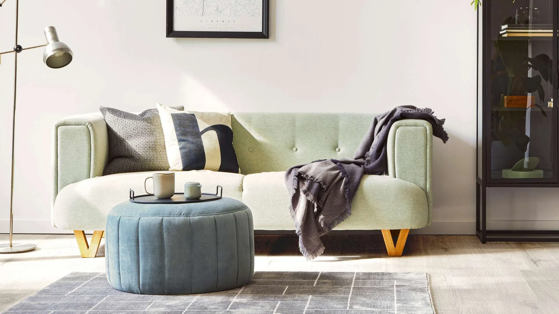 Modern living room with light green sofa, blue ottoman, cozy throw, and floor lamp on wooden floor.