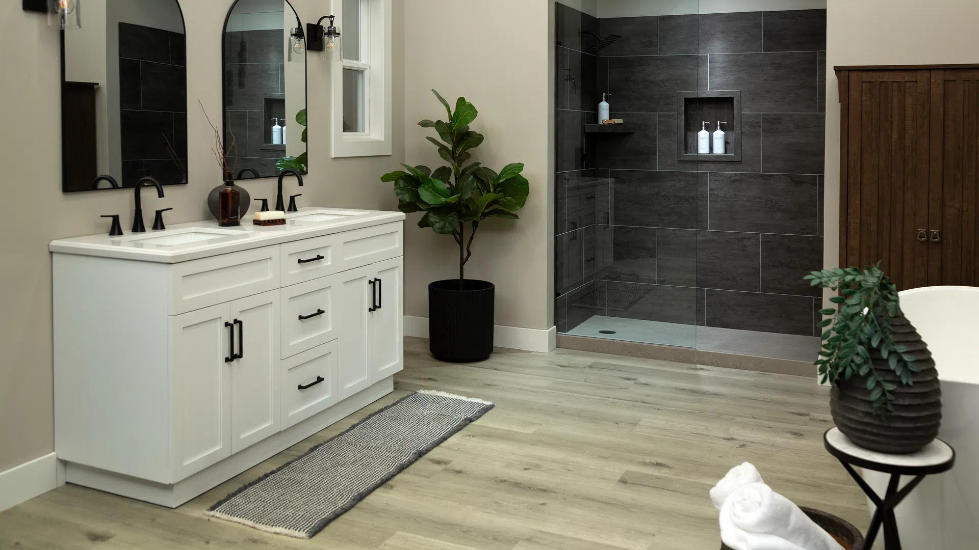 Modern bathroom with white vanity, black fixtures, large mirrors, walk-in shower, and wood flooring.