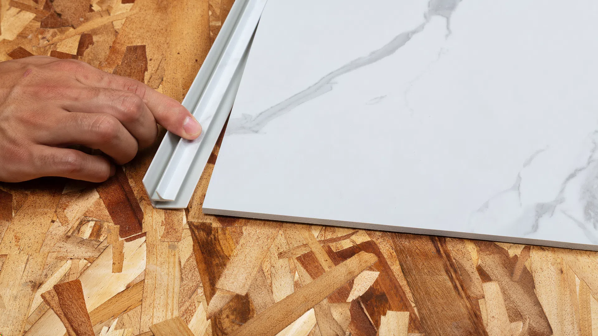 Hands installing white marble-patterned tile with plastic trim on plywood subfloor surface.