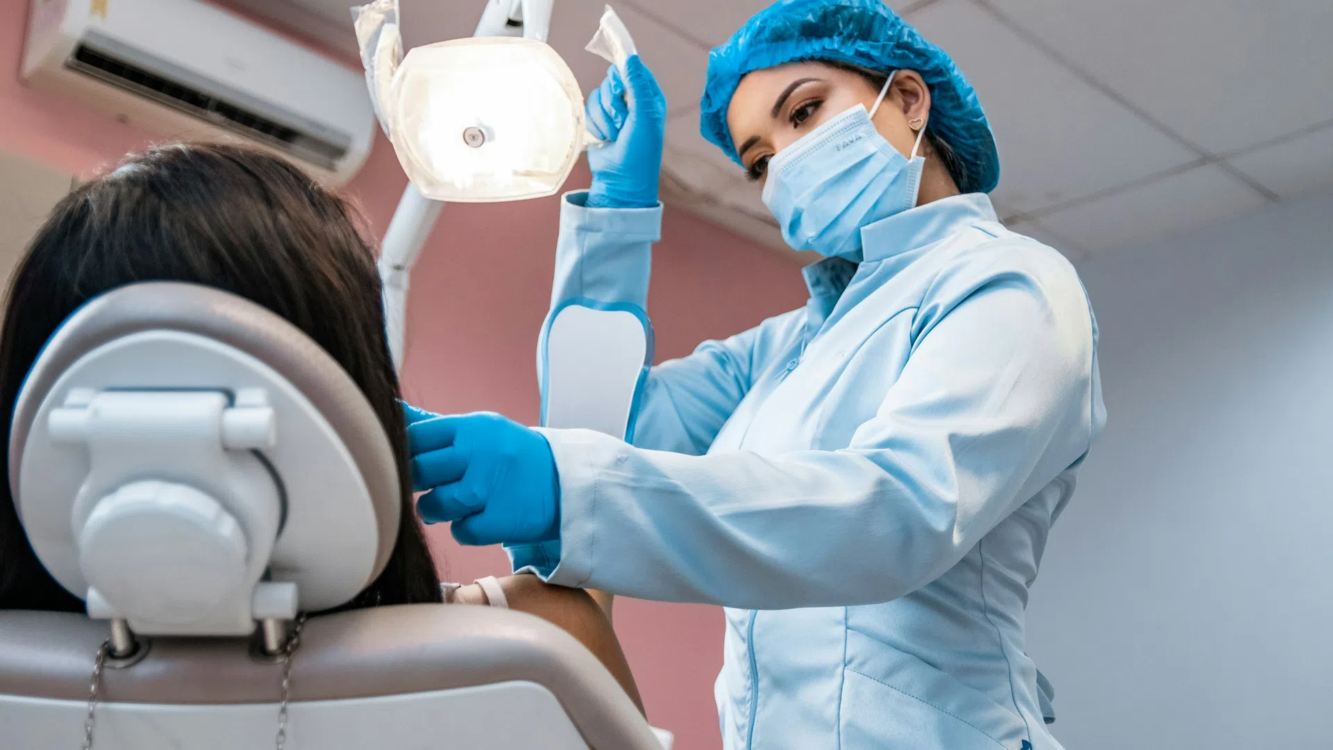 Female dentist in protective gear adjusting overhead dental light for patient in clinic chair
