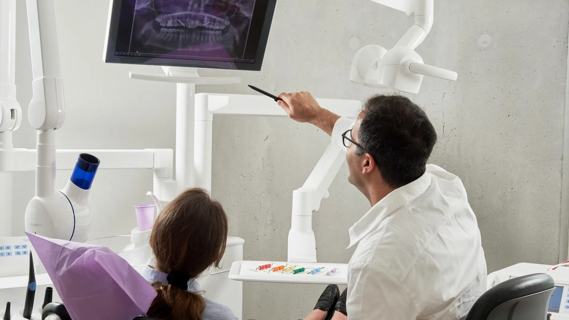 Dentist explaining dental X-ray results to patient seated in clinic chair under bright light.