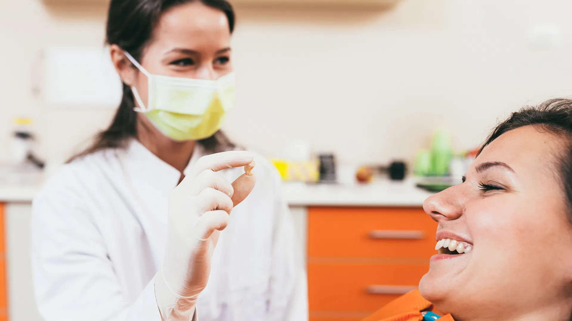 Dentist wearing mask and gloves holding a dental crown while smiling patient reclines in dental chair