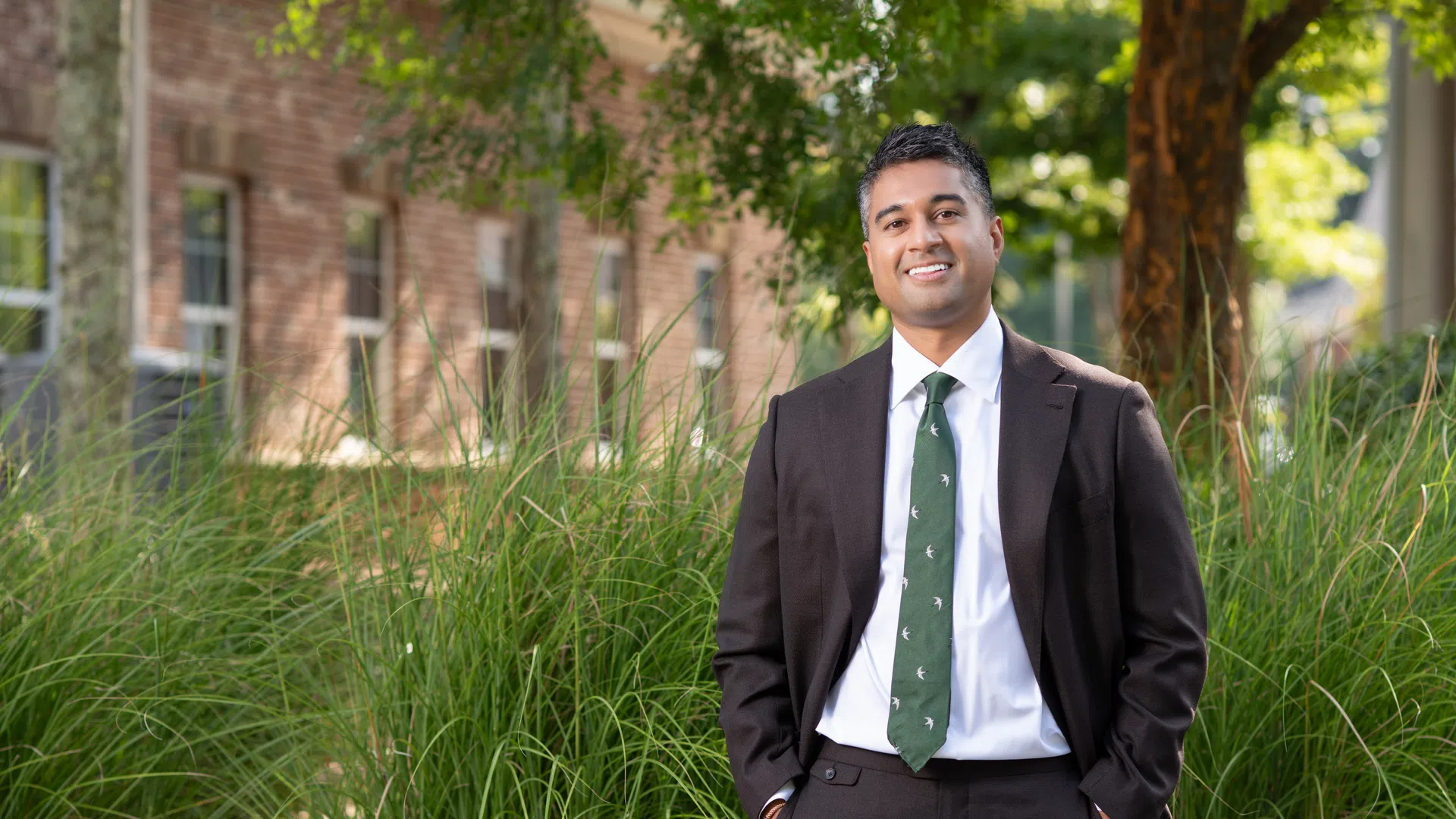 Smiling man in dark suit and green tie standing outdoors in front of tall grass and trees with building background