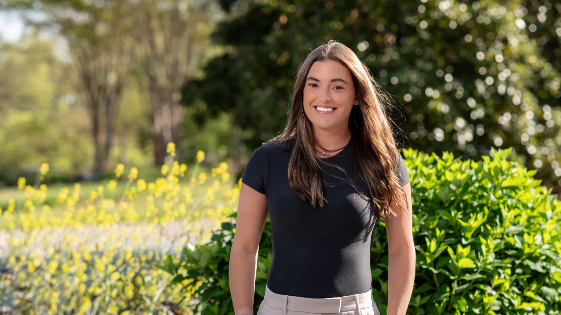 Smiling young woman in black shirt and beige pants standing outdoors with greenery and yellow flowers in background