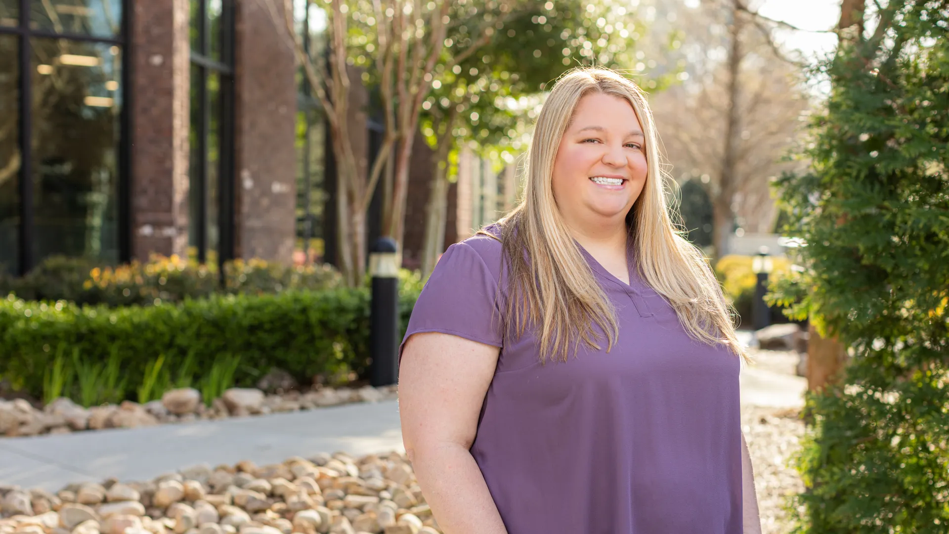 Smiling woman in a purple shirt standing outdoors near rocks and greenery on a sunny day.