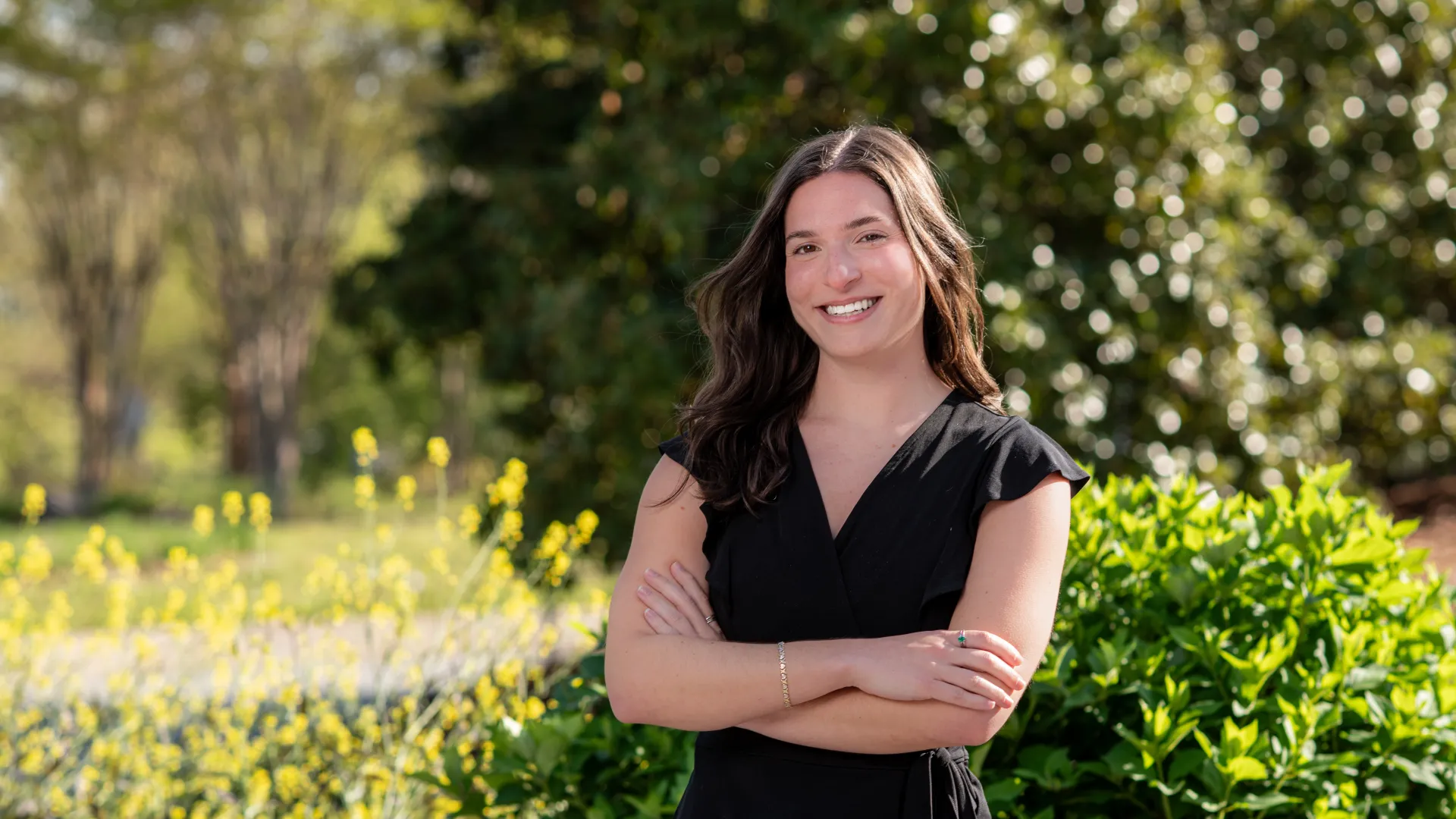 Smiling woman in black dress standing with arms crossed in sunny garden with greenery and yellow flowers.