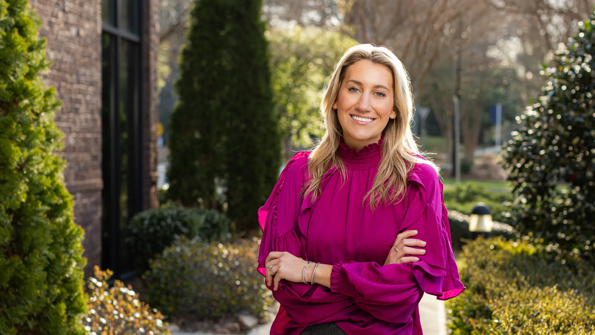 Smiling woman in a magenta blouse standing outdoors with arms crossed against a backdrop of greenery and buildings.