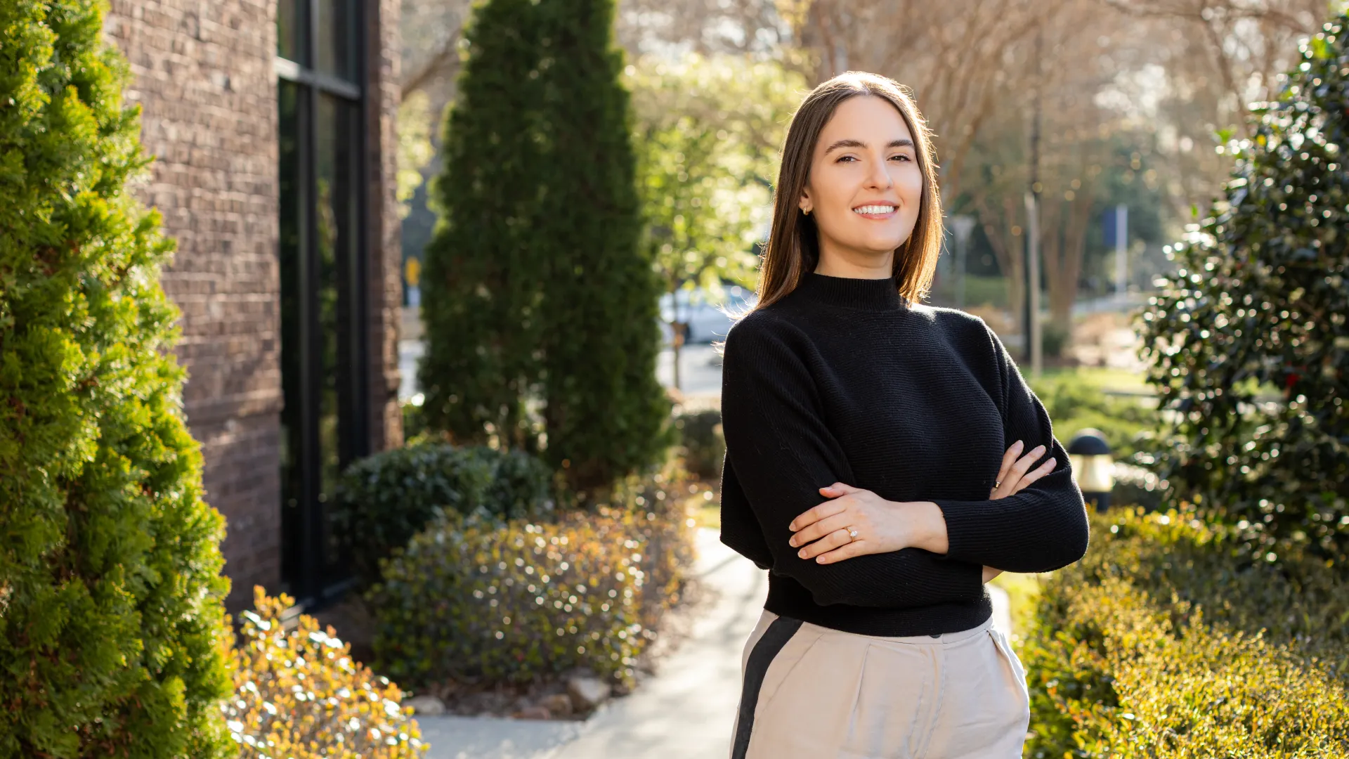 Confident young woman in black sweater standing outdoors with crossed arms and smiling in sunlight.