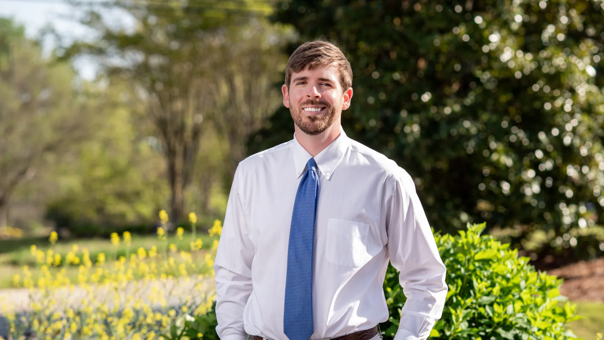 Smiling young man in white shirt and blue tie stands outdoors with green bushes and trees in background.