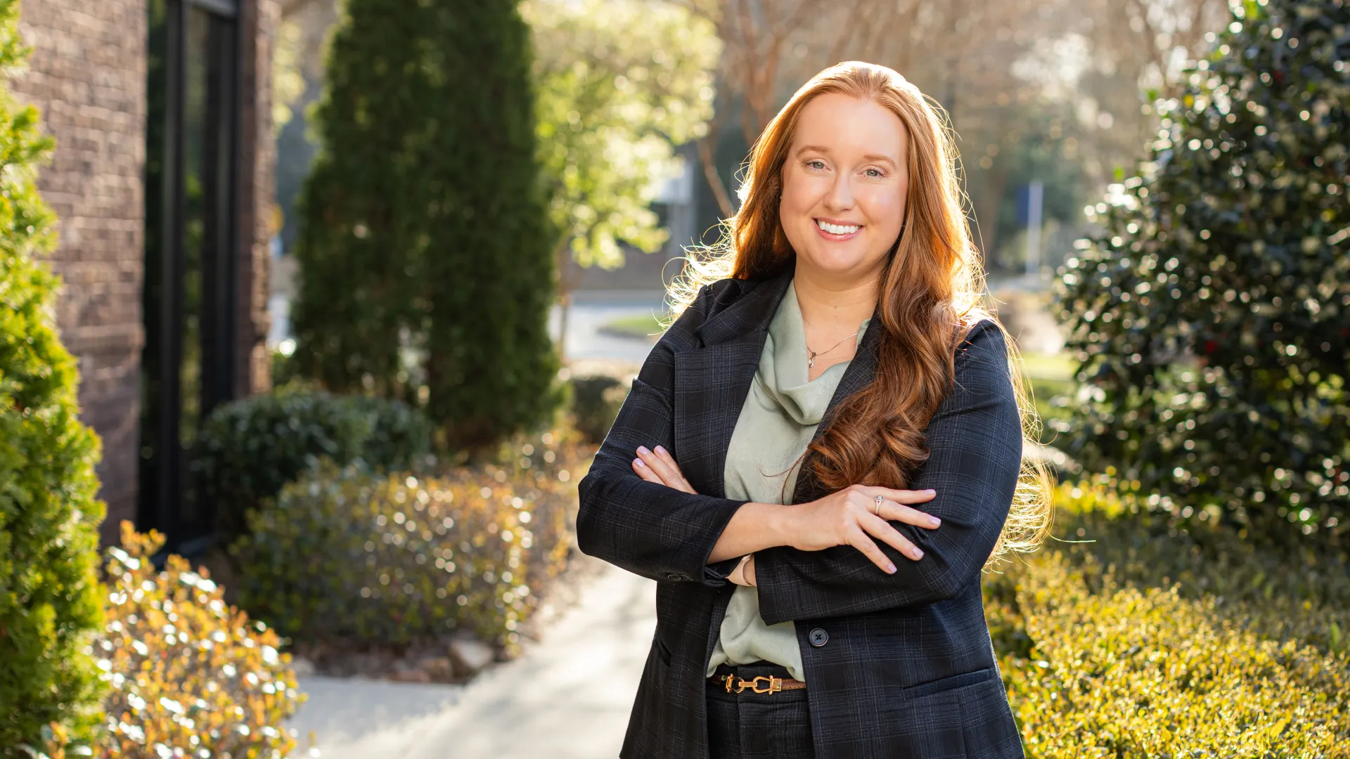 Confident woman with long red hair in a dark suit standing outdoors with arms crossed on a sunny day.