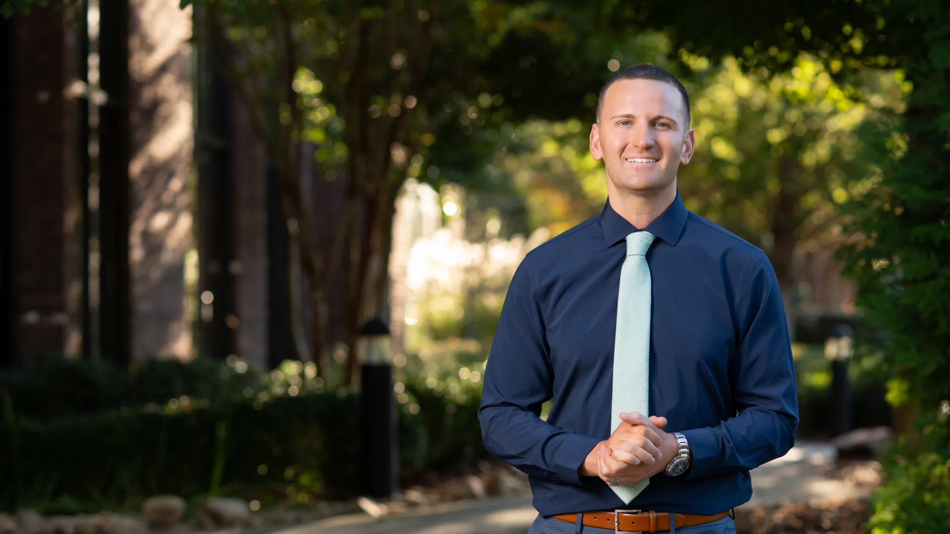 Man in navy shirt and light tie standing outdoors on a sunny path with trees and greenery in background