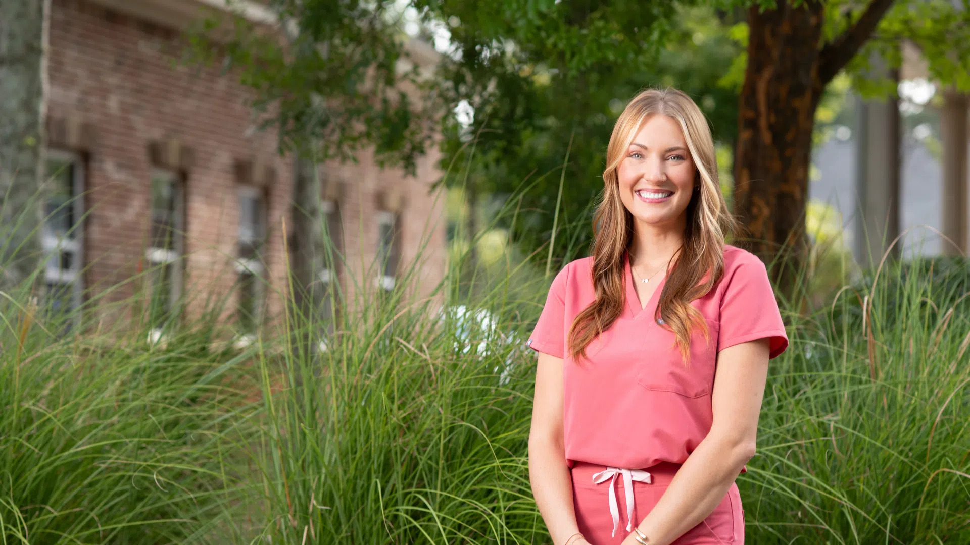 Smiling woman in pink scrubs standing outdoors with green grass and trees in the background on a sunny day