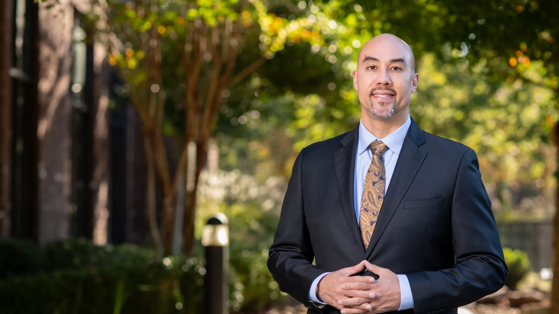 Confident bald man in black suit and patterned tie standing outdoors with blurred trees and building in background