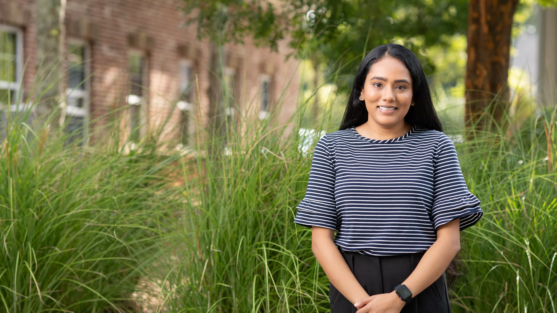 Young woman in a striped shirt standing outdoors with tall grass and brick buildings in the background
