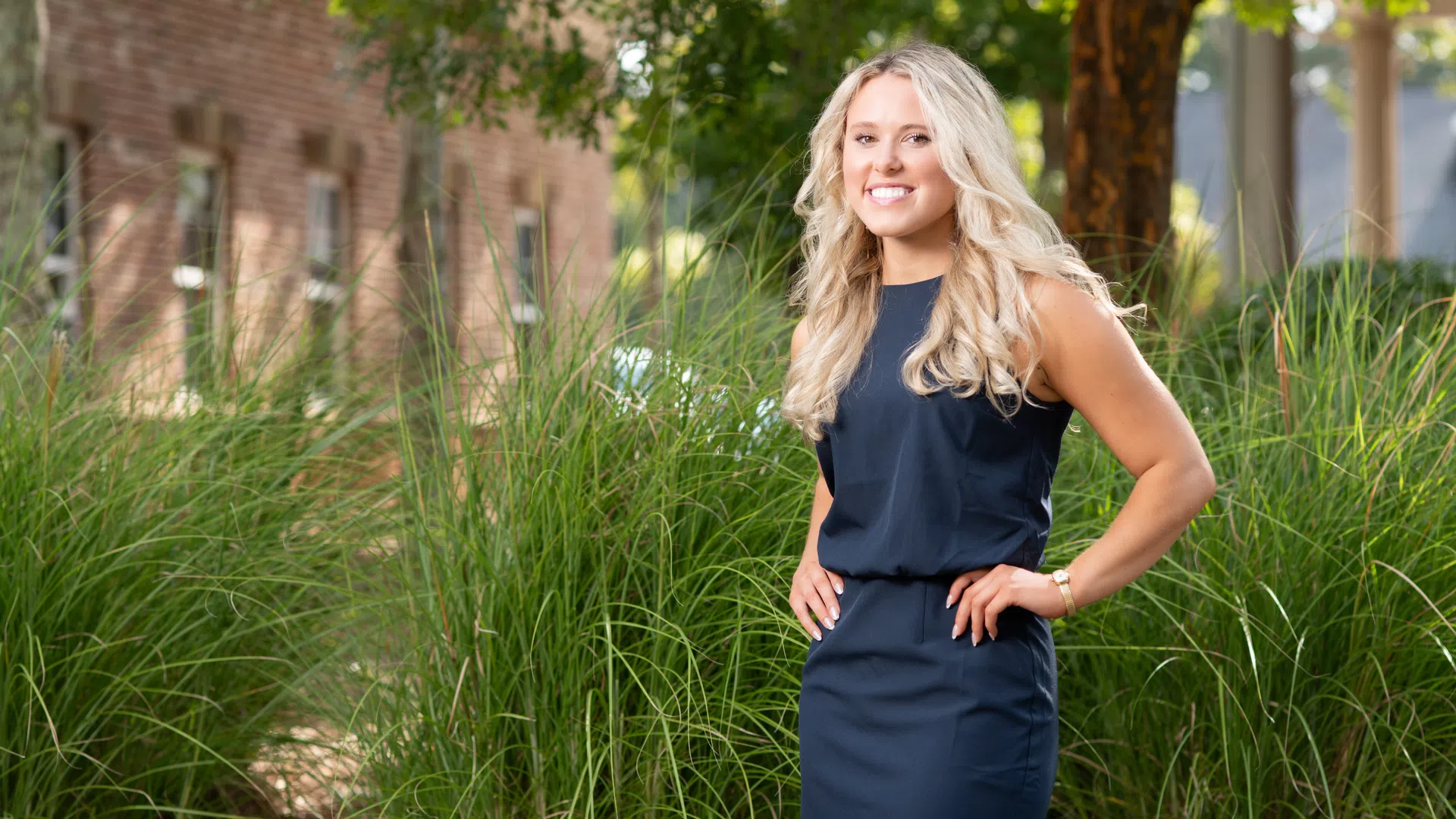 Smiling blonde woman in a navy dress standing outdoors among tall green grass with trees and a building in the background