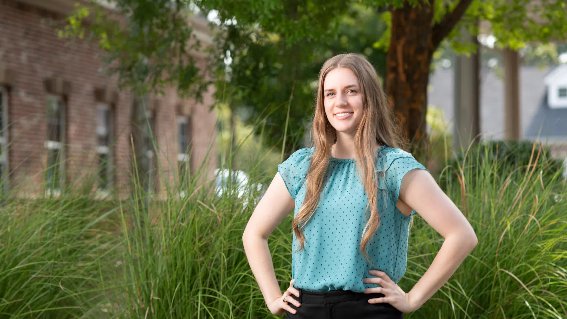 Smiling woman in teal blouse and black pants standing confidently outdoors with hands on hips near green plants.