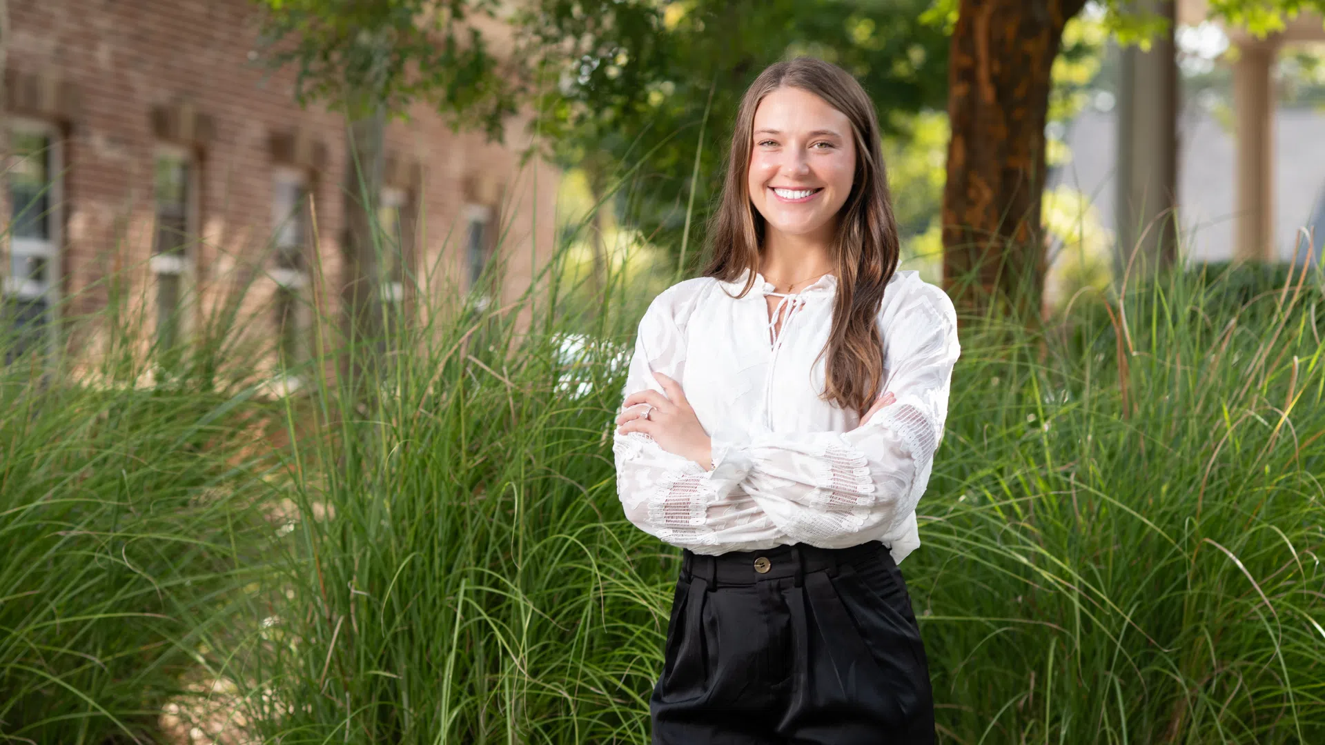 Smiling young woman in white blouse and black pants standing outdoors with green grass and trees in background