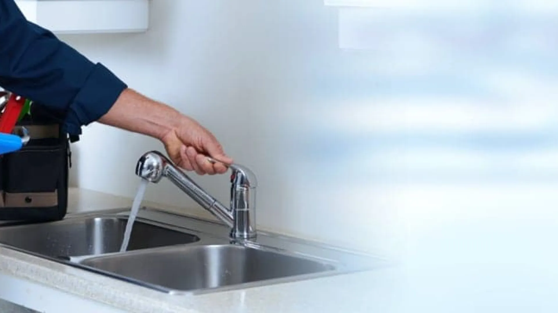 Plumber checking water flow from kitchen faucet with clipboard and tool bag nearby in modern kitchen sink area.