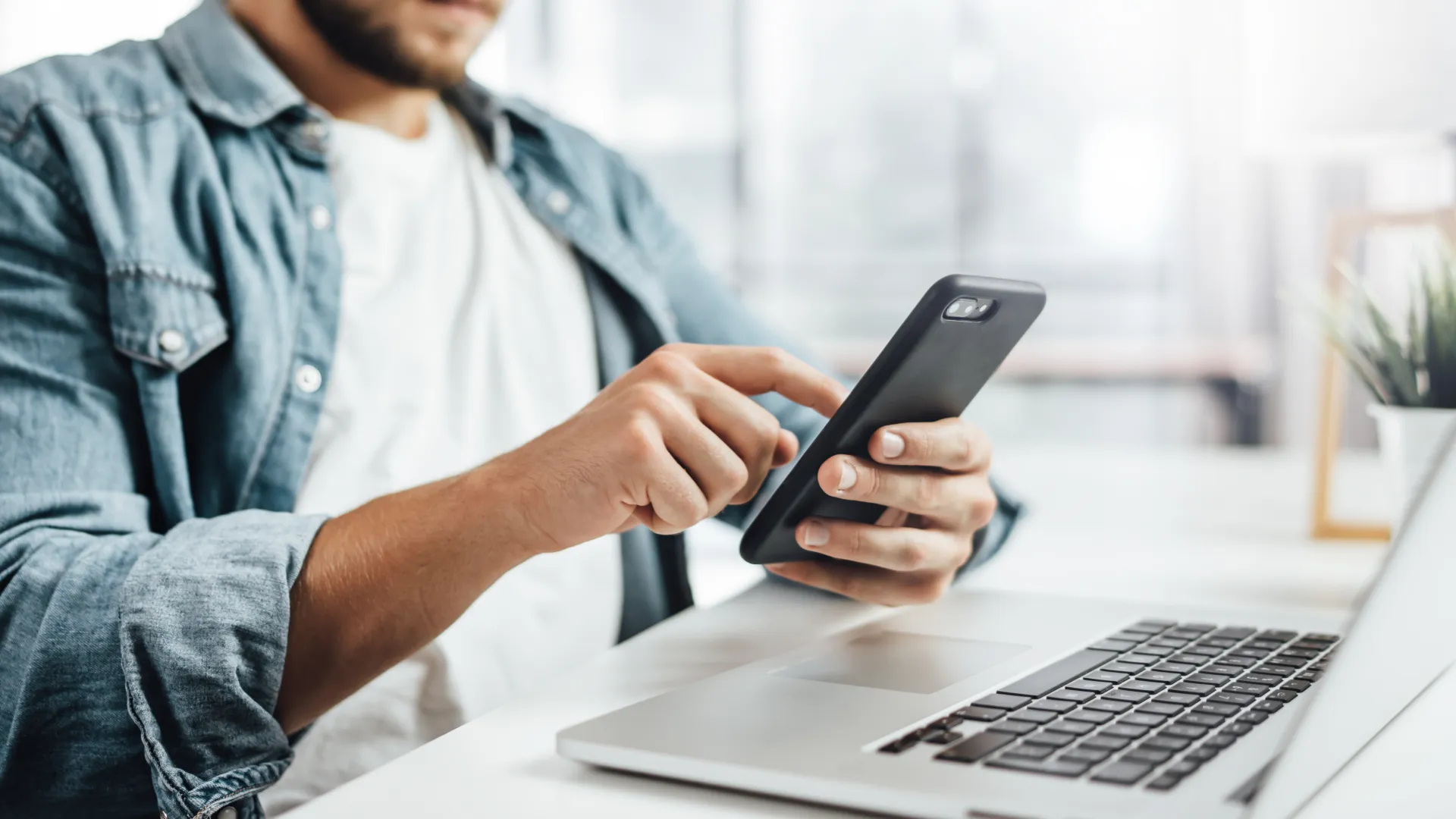 Man using smartphone near laptop on desk in a bright modern workspace with casual attire