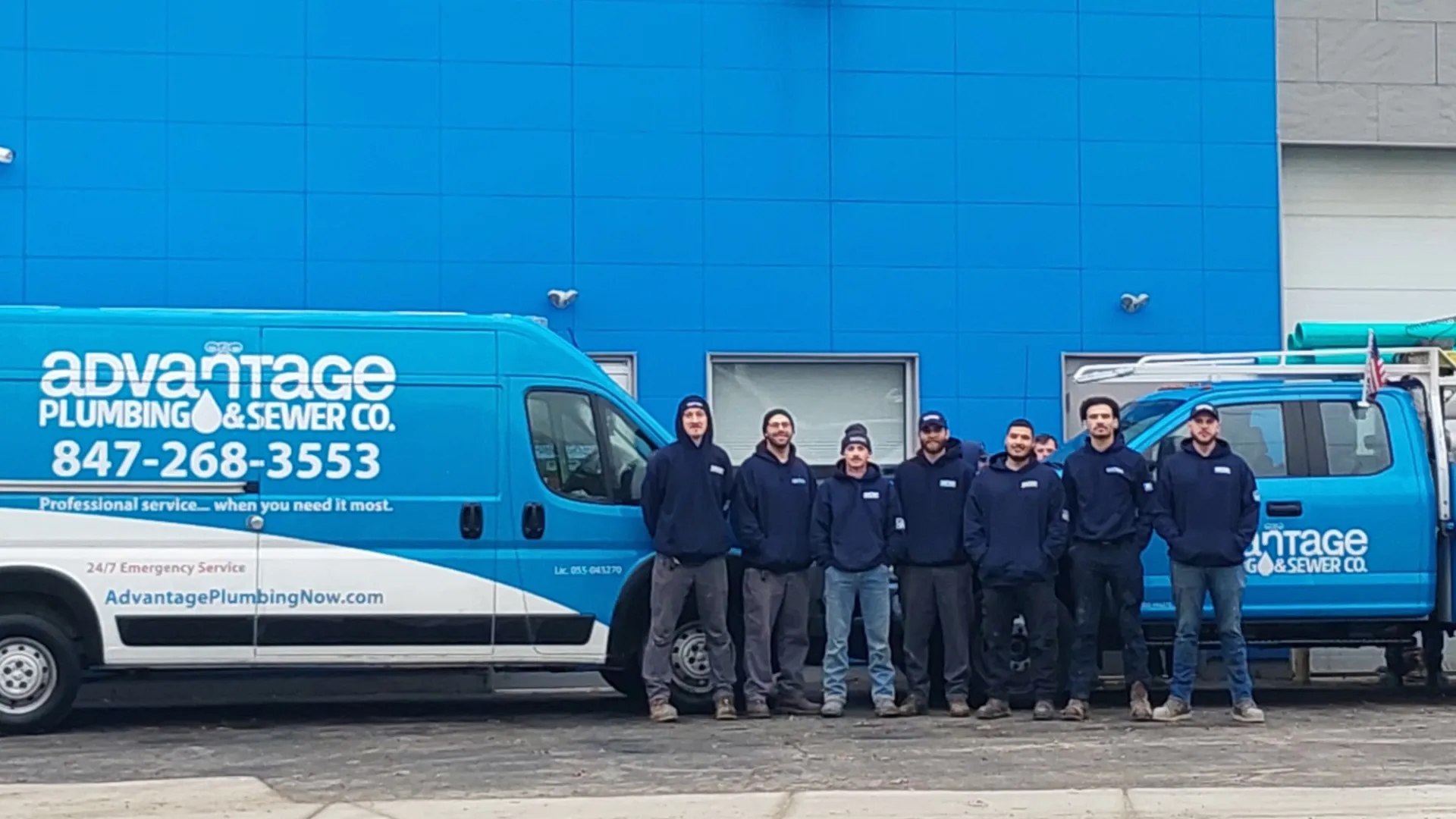 Team of plumbers standing in front of two blue Advantage Plumbing & Sewer company vehicles outside a blue building.