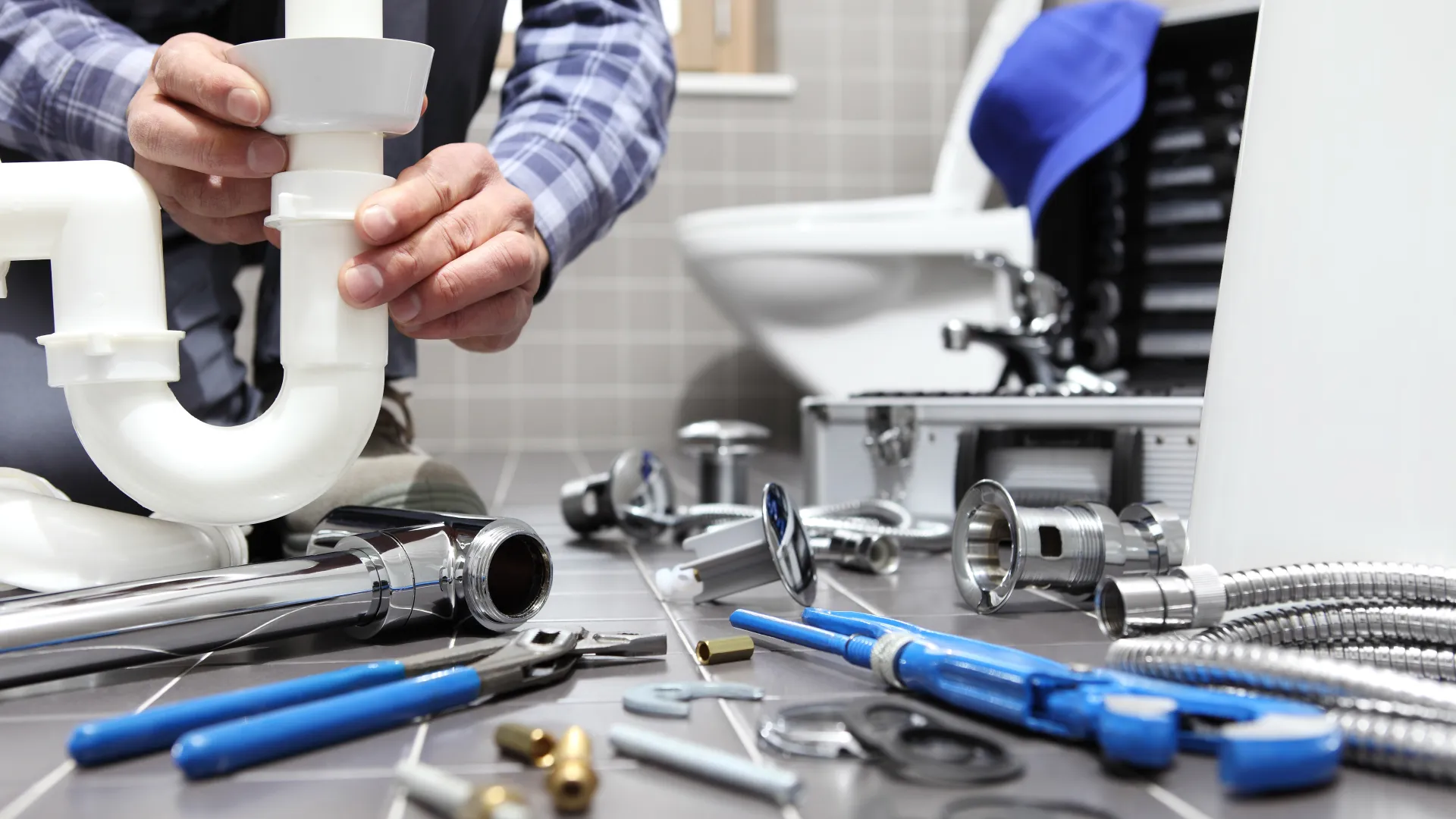 Plumber installing white PVC pipe under bathroom sink surrounded by tools and metal plumbing parts on tiled floor