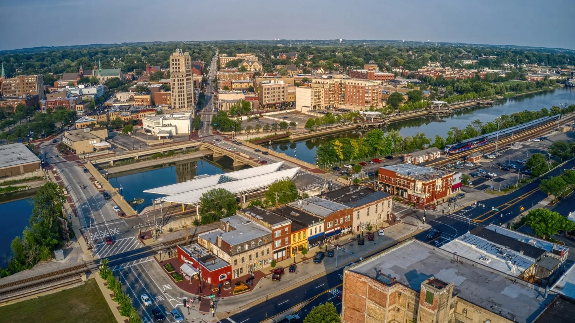 Aerial view of a riverfront city with bridges, historic buildings, train tracks, and green trees under a blue sky.