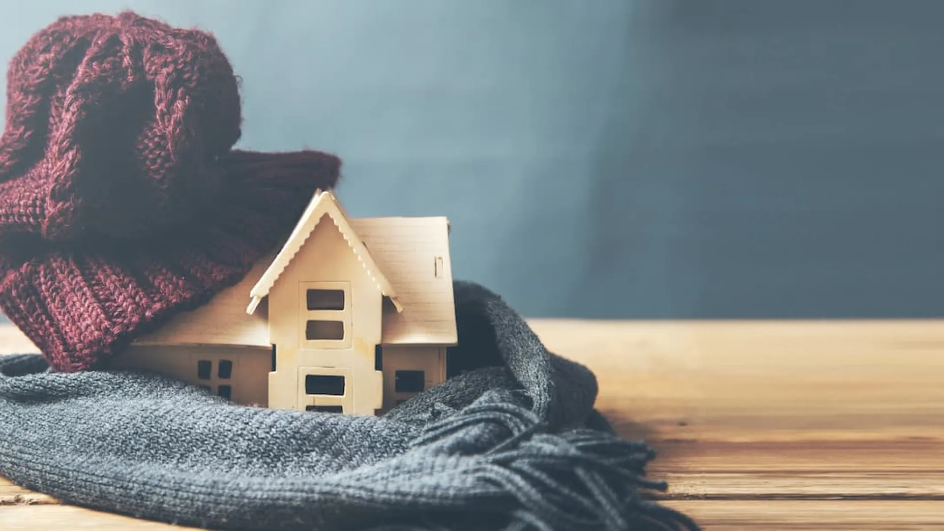 Miniature wooden house wrapped in cozy scarves and a knit hat on a wooden table with gray background