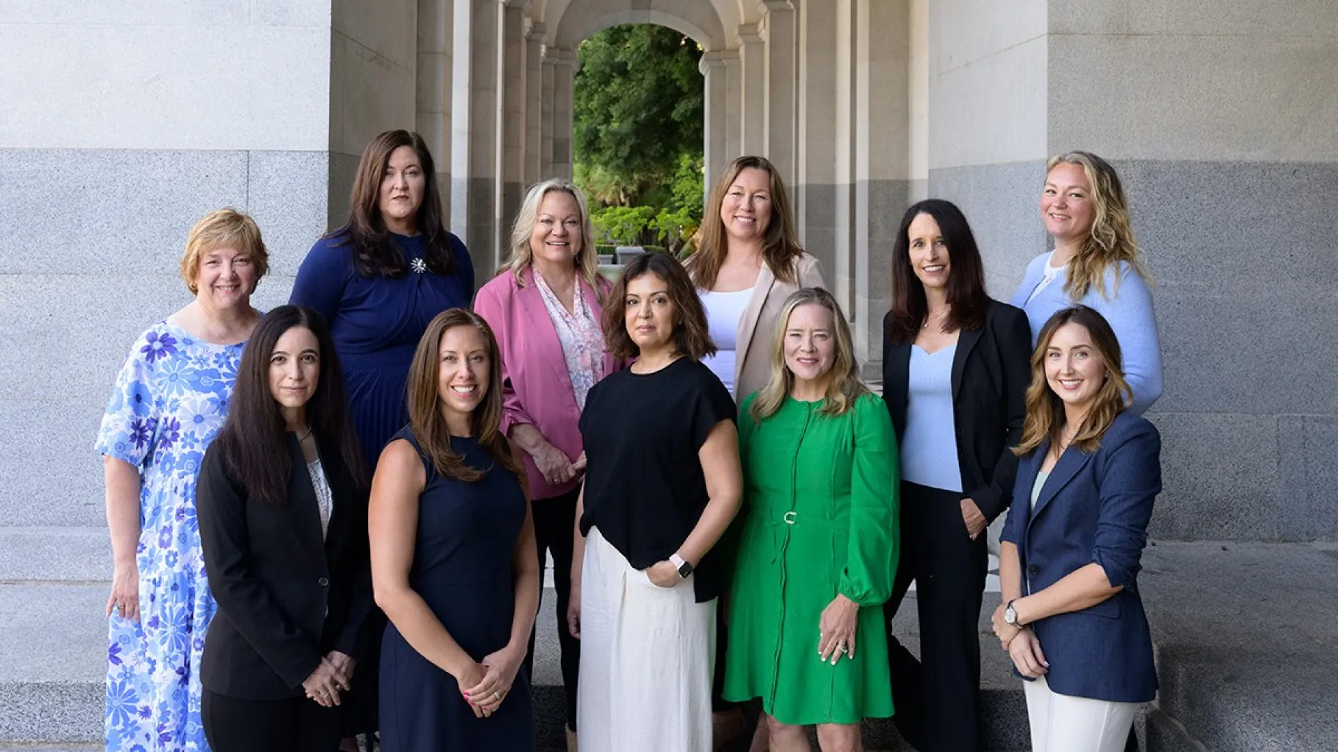 Group of professional women standing on stone steps under an archway with greenery in the background.