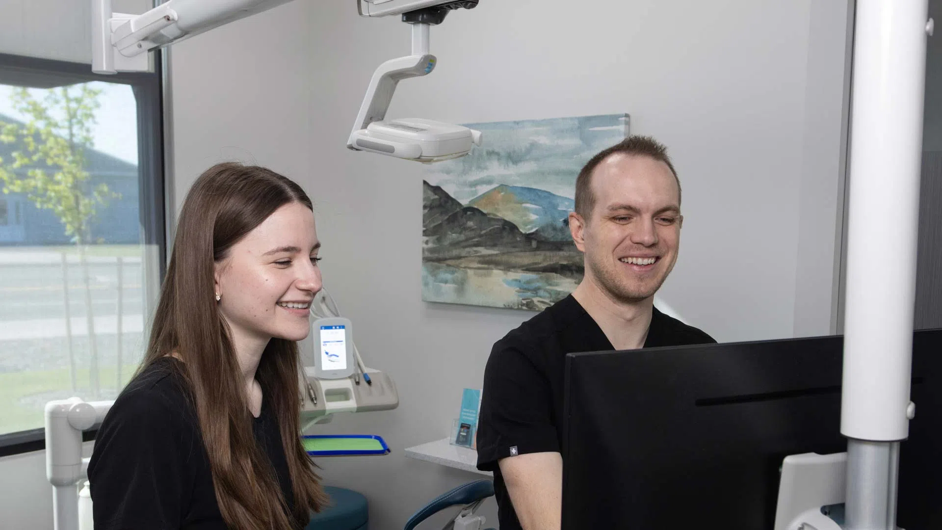 Two dental professionals in black scrubs smiling while looking at a computer screen in a clinic.