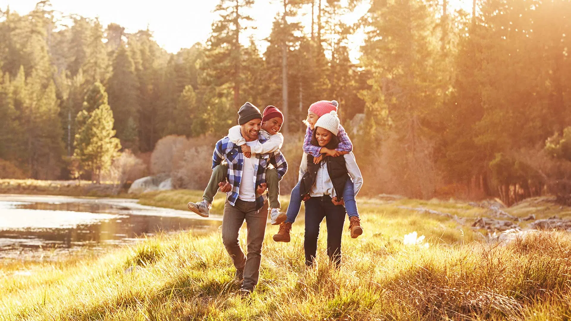 Family enjoying a sunny autumn day outdoors with children riding piggyback near a lake and forest.