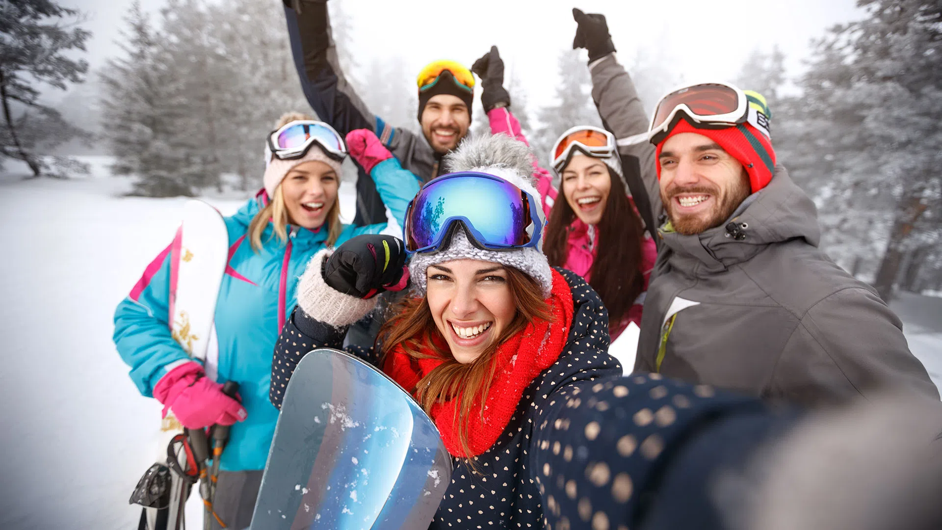 Group of five friends in colorful winter gear taking a selfie while snowboarding in snowy forest.