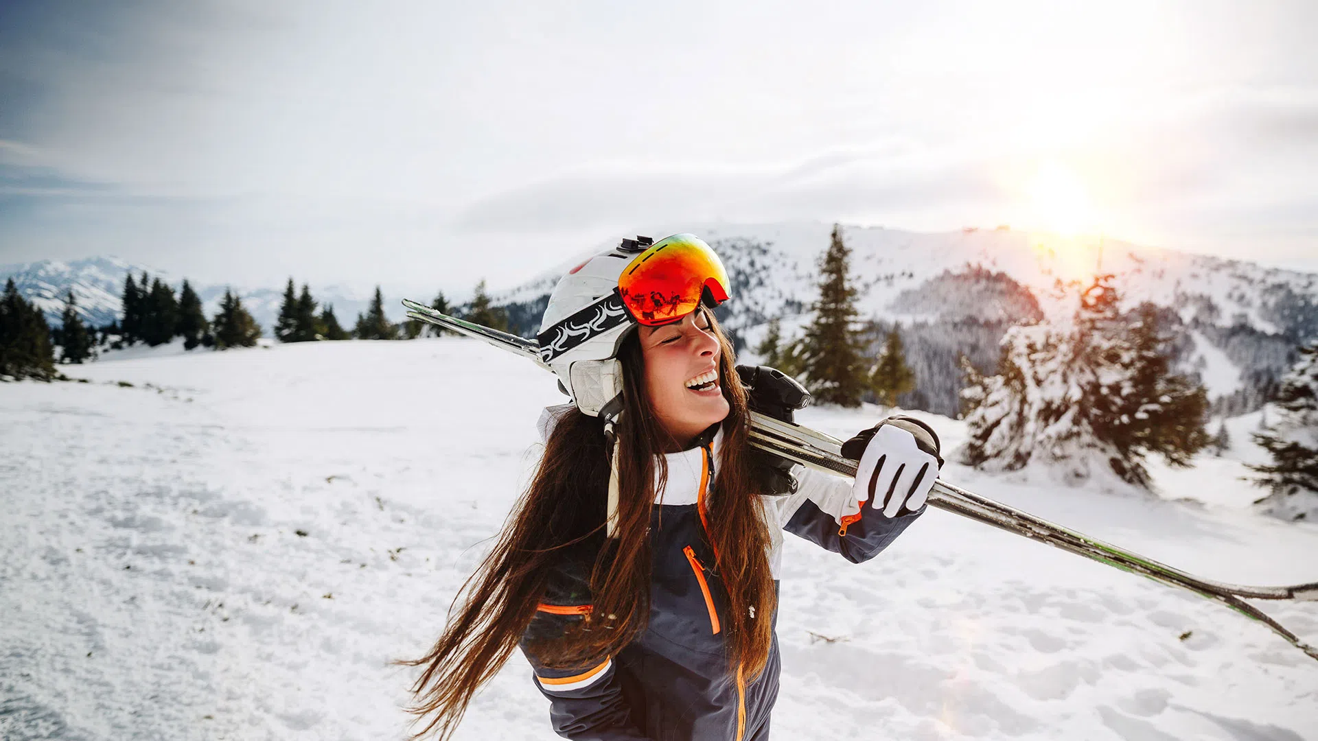 Smiling woman skier carrying skis on snowy mountain landscape at sunset with pine trees in background