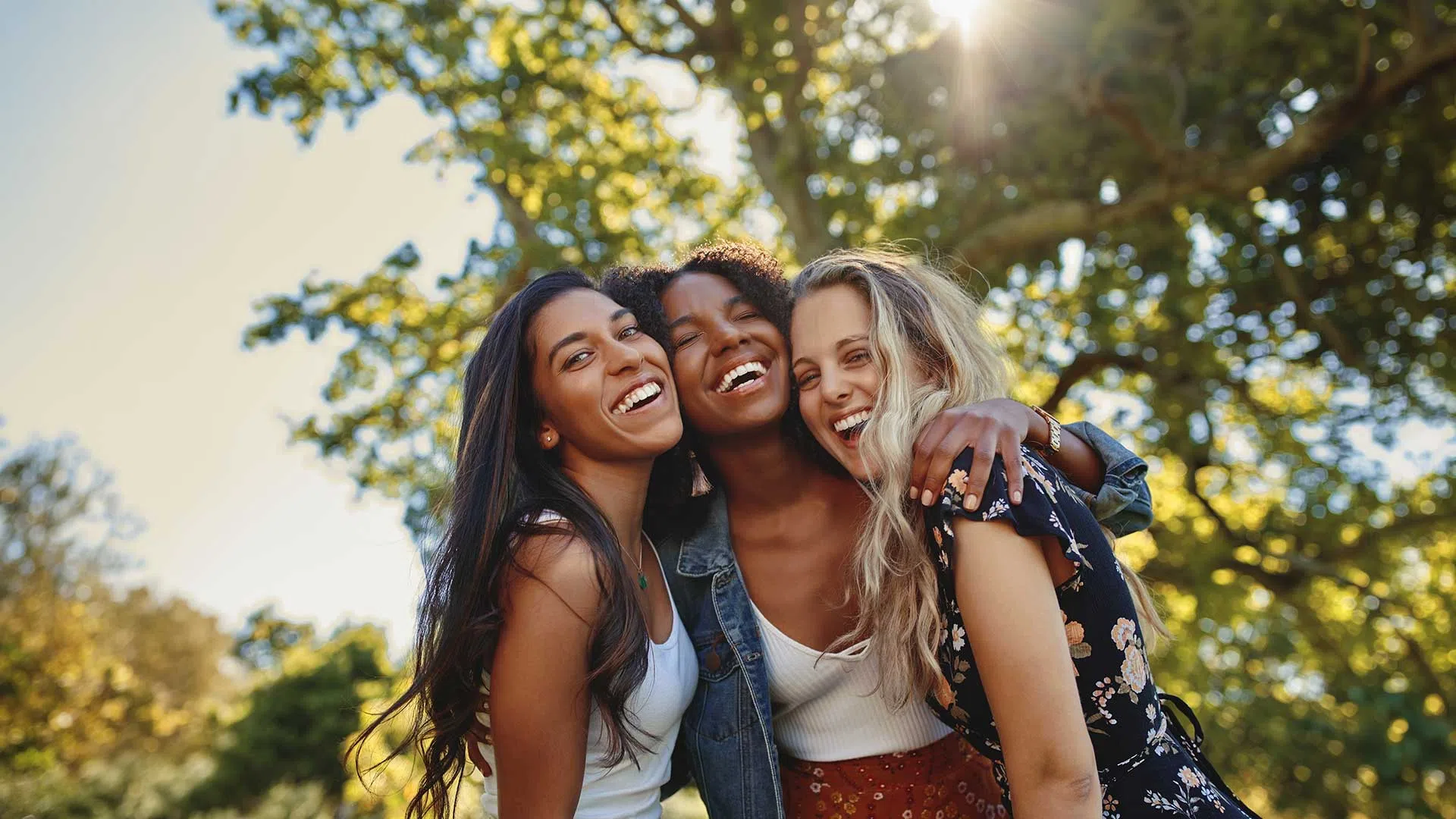 Three diverse women smiling and hugging outdoors with sunlight and trees in the background