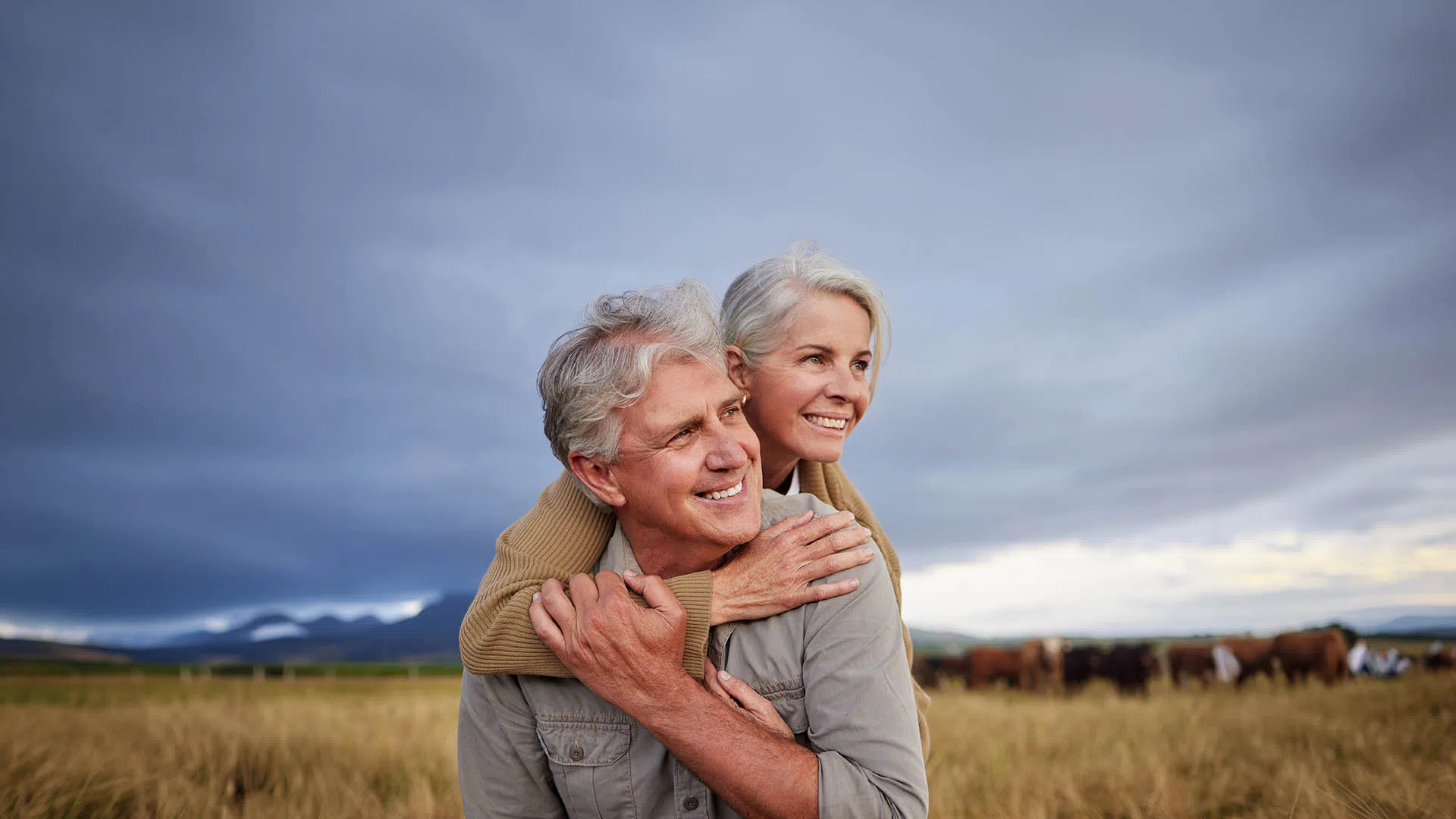 Smiling senior couple enjoying a countryside view with cattle and mountains under a cloudy sky.