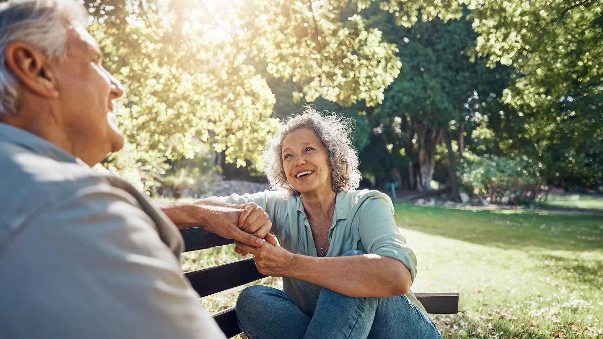 Smiling mature woman holding hands and sitting on park bench with elderly man in sunny green garden.