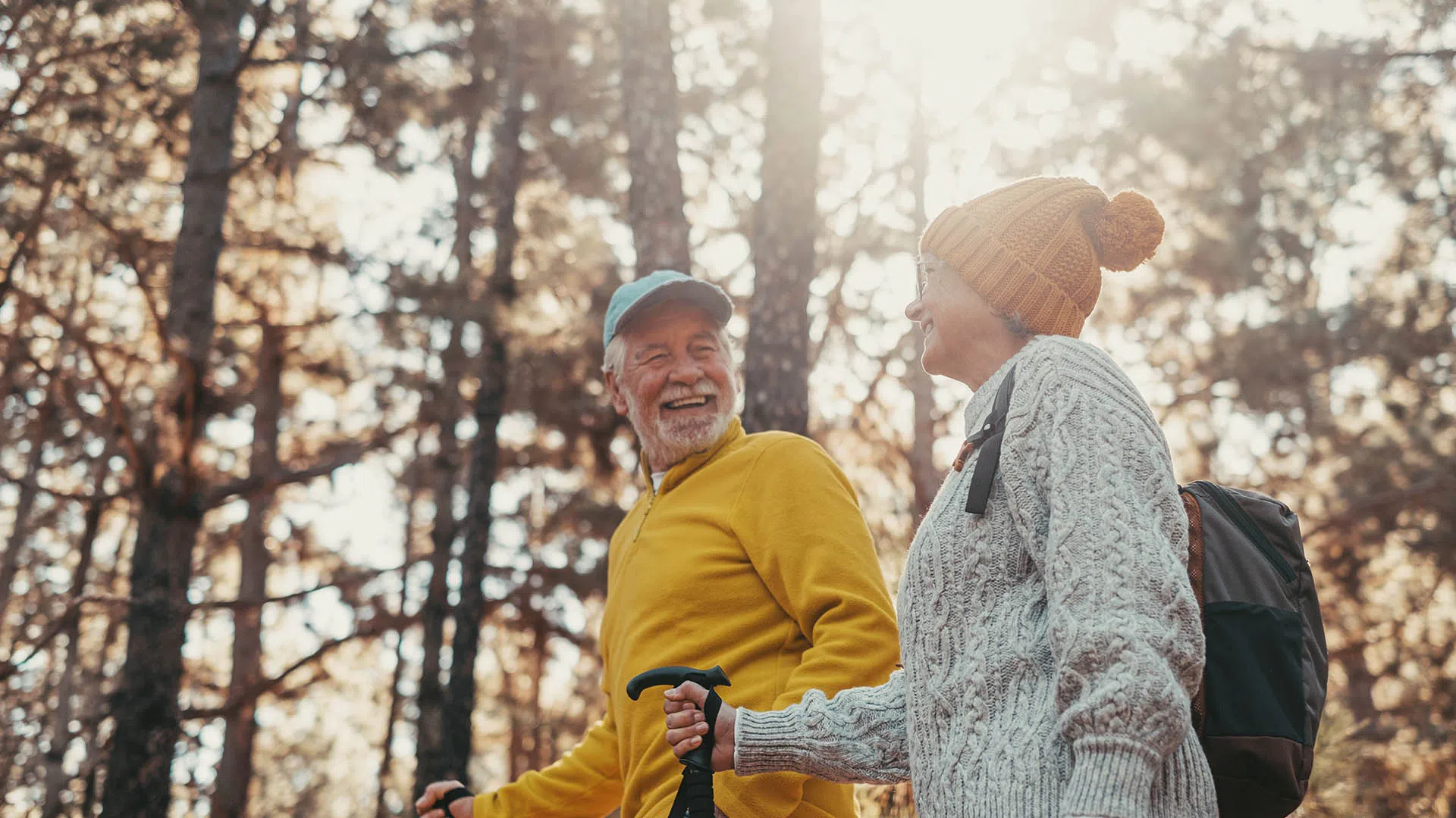 Smiling elderly couple hiking in autumn forest wearing sweaters and hats with walking sticks.