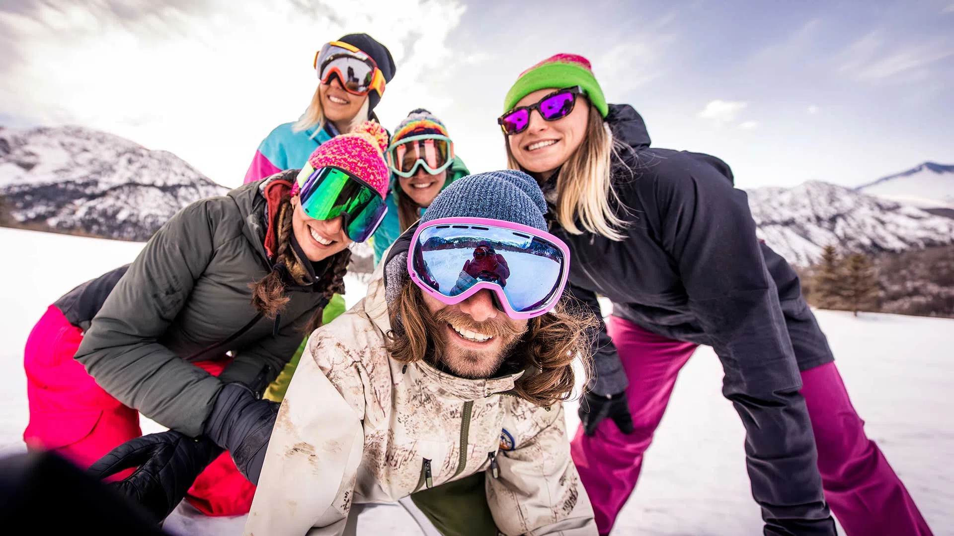 Group of five friends wearing colorful ski gear smiling and posing on snowy mountain slope under clear sky.