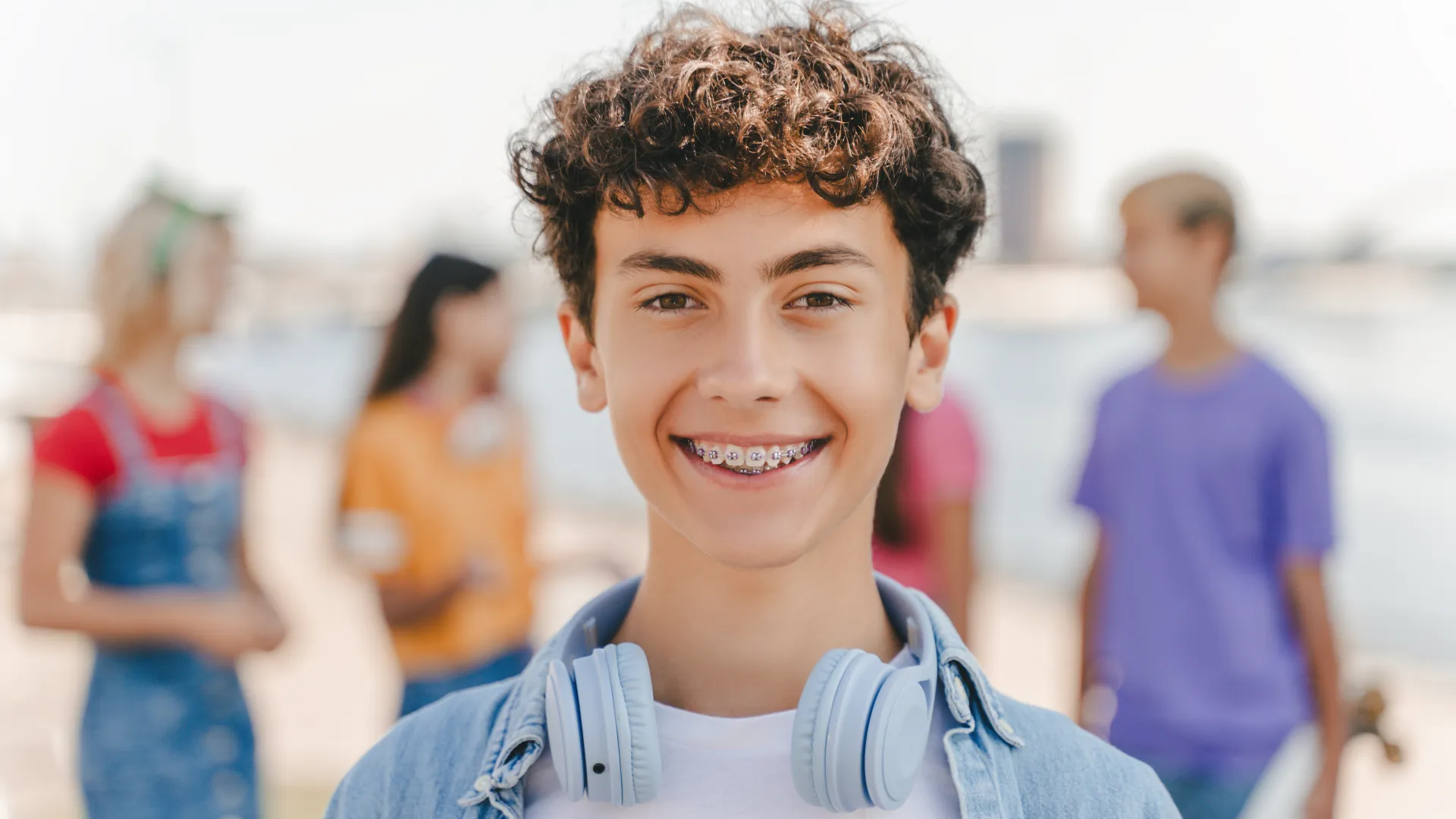 Smiling teen boy with curly hair and braces wearing headphones around neck with friends outdoors.