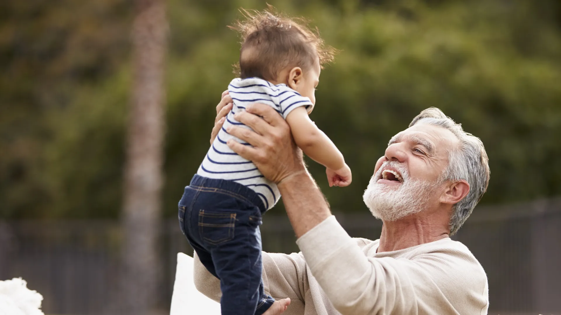 Smiling elderly man lifting a baby boy outdoors, showing joy and bonding in a natural setting.